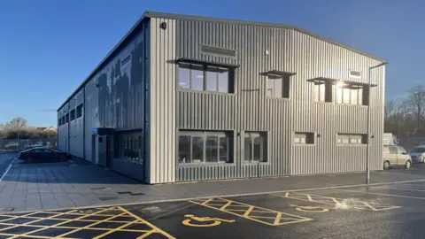 The exterior of the new centre in Bideford. It is modern and has grey panels. There are large windows around it and a parked car to the left of it. In front of the building there are disabled parking bays.