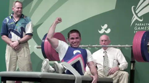 Anthony Peddle Smiling man lying on a gym bench with his arm in the air