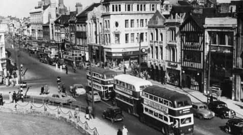 A black and white photo of Cardiff city centre. There are lines of shops on one side, castle green on the other. Through the middle is a road with double decker buses and cars on it and there are lots of people milling about.
