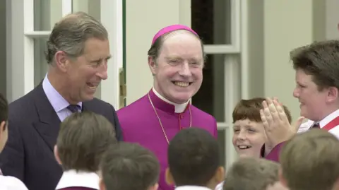 King Charles III (then a prince) standing next to Bishop McMahon near a doorway. He is wearing a dark suit and smiling as he speaks to choirboys. Bishop McMahon is dressed in a bright magenta clerical robe with a matching skullcap.