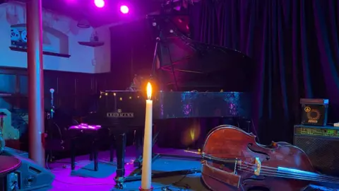 A grand piano on the Blue Lamp stage in front of curtain. In front of it lies a violin on a table and a candle. 