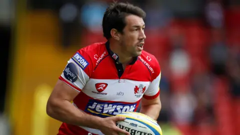 Action Images Tom Voyce playing for Gloucester, wearing a reddish  and achromatic  garment  and carrying a ball.