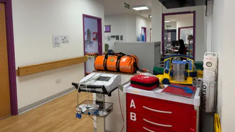 A corridor at the Urgent Treatment Centre at Woking Community Hospital. Medical equipment and a red chest of drawers can be seen. In the background, there is a receptionist.