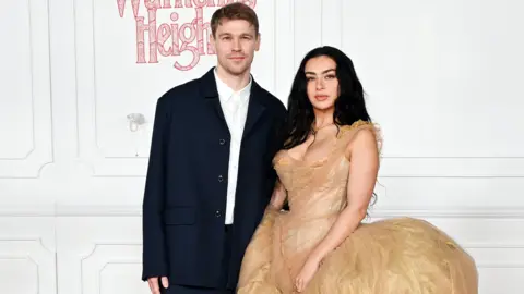 Finn Keane and Charli XCX standing next to each other at the Wuthering Heights premiere. There is a white wall behind them. Finn is wearing a white shirt with a dark navy jacket unbuttoned and is staring directly at the camera. Charli has her long black hair down and is wearing a gold dress.