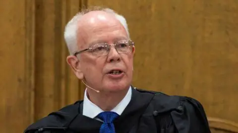 Getty Images Jim Wallace is standing before the wooden walls of the Assembly Hall in Edinburgh. He is wearing a black ceremonial robe, fastened at his collar, black suit, white shirt and mid-blue tie. 