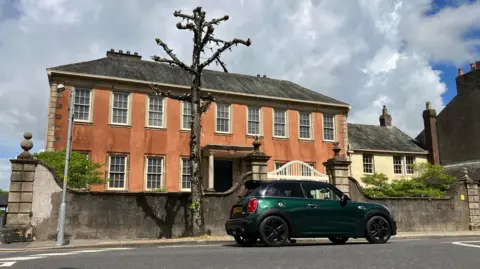 A pollarded tree outside Wordsworth House on Main Street in Cockermouth. The brown rendered Georgian town house is over two levels and has sash windows all along the facade. There is a large gate at the front, with a green mini driving in front of it.