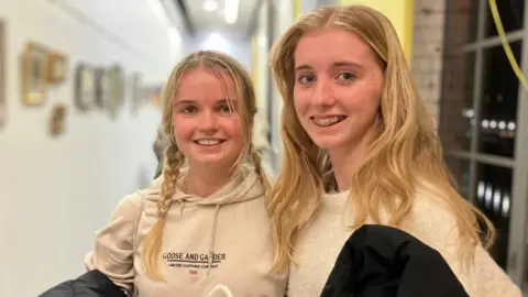 BBC/Seb Cheer Two young women with blonde hair smile at the camera, standing in a corridor.