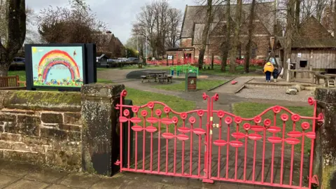 A small park with a red gate at the front and a rainbow sign with benches and play equipment and trees in the background and a large sandstone building