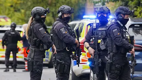 PA Media Armed police officers at the scene near the Heaton Park Hebrew Congregation synagogue in Crumpsall, Manchester, where two people died in a suspected terror attack by a man who was shot dead by police. The four officers are dressed in black, holding assault rifles and wearing helmets with face coverings drawn up over their mouths. 