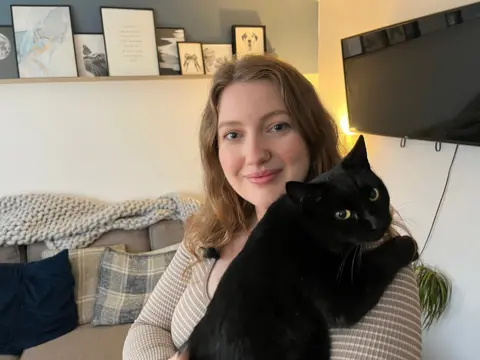 A woman with long brown hair holding her black cat looking at the camera