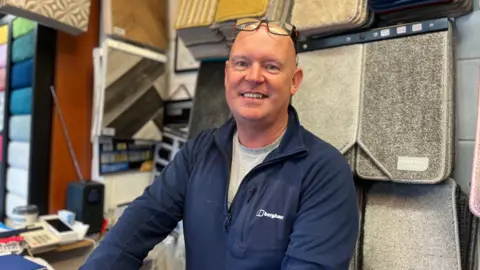 Shop owner Ben Corrie, who is bald and has glasses on his head, is wearing a navy fleece over a grey t-shirt. He's behind a desk in a shop with samples of carpets handing on a shelf behind him.