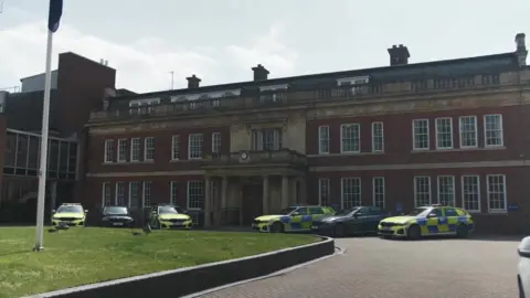 Kate Bradbrook/BBC A three-storey brick building with rows of windows and six police cars parked outside.