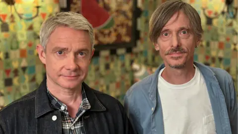 Two men stood in front of a colourful wall looking into the camera