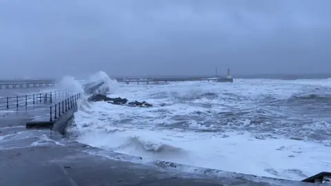 BBC Weather Watchers / Susan White, frothy waves are crashing over the pier at Amble. The sea is foamy and choppy. There is a white-and-red lighthouse in the distance.
