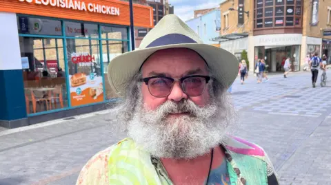 Phil Harrison A man wearing glasses with a big white beard, a colourful shirt and sun hat. He is standing at a high street.