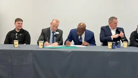 Ollie Conopo/BBC Four men sit at a table covered in a grey table cloth. Frank Bruno, in a blue suit, signs a document with former MP Chris Heaton-Harris, wearing a charcoal suit.