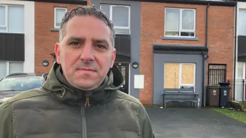 Christopher Jackson has short hair and is wearing a large green coat. He is standing near a house.