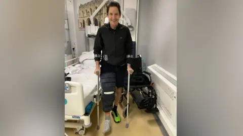 Andrew Rawlings in a hospital ward standing on crutches beside a hospital bed, with a wheelchair behind him