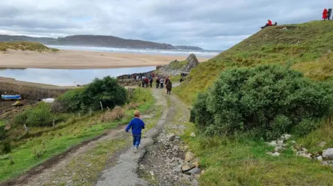 Strathnaver Museum A boy runs down a track towards Bettyhill pier for the launch of the new boat. There is a crowd of people - and the beach - ahead of him.