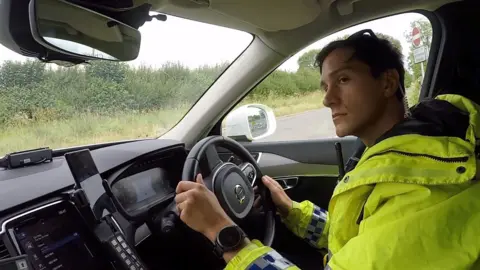 An officer in a high-vis yellow jacket at the wheel of a vehicle. He is looking to the left and has both hands on the wheel, and an earpiece in his left ear. He appears to be by a junction, with a no entry sign to his right.