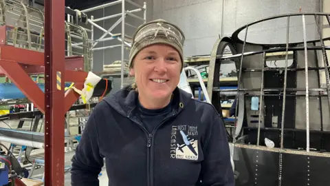 BBC/Karl Bird Volunteer Jodie stood in a warehouse smiling at the camera. She is wearing a khaki beanie and work glasses on her head. She is also wearing a navy fleece and a navy t-shirt. 