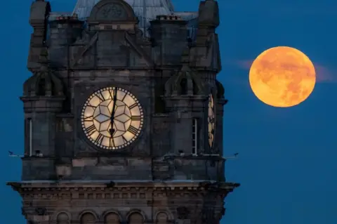 PA Una súper luna azul, que parece de color naranja brillante, junto al Reloj Balmoral en Edimburgo en agosto pasado