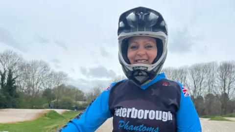 Connor Bennett/BBC Leah Pearson, a woman wearing a BMX helmet. She is looking directly at the camera and is smiling. She is standing on a BMX track which trails off behind her.