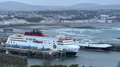 BBC The Manxman and Manannan ferries, which are both white, red and black, moored in Douglas Harbour. Choppy seas in Douglas Bay are in the background.