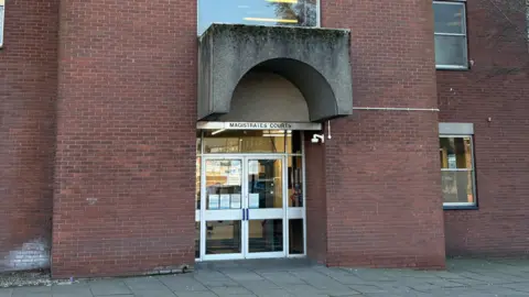 The entrance to Ipswich Magistrates' Court. Brown brick walls can be seen either side of a glass doorway.