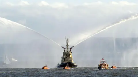 RNLI/Tom Collins A large white and blue boat spraying water out of its sides. Smaller orange boats surround it.