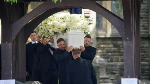 PA Media A group of pallbearers carry a white coffin with an angel wings design on the end. The coffin is adorned with white roses and lilies. The group are about to go through a gateway at the church.