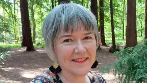 Melanie Johnson-Remy A woman with short grey hair, black and gold earrings, and a multi-coloured top, smiles as she takes a selfie in the middle of a wooded area.