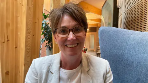 A woman wearing a cream suit jacket and white top is sat on a blue chair, smiling.