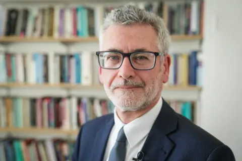 Prof Shane O'Neill is wearing a suit and tie. He has grey hair and black glasses and is standing in an office with a bookcase behind him.