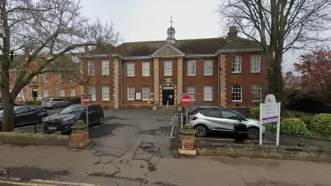A front view of the Fenland District Council building. It is redbrick with sash windows and a large entrance door with steps leading up to it. There is a tower on top of the building and cars are parked in a car park area at the front