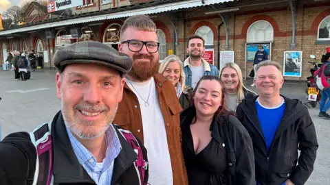 Supplied A man takes a selfie style image with several other adults behind him. They are all smiling at the camera. They are standing in front of Ipswich train station.