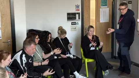Jodie Halford/BBC Six people sit on chairs and look at their finger tips, while a man stands next to them and instructs them on how to deliver first aid.