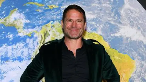 Steve Backshall a man who is wearing a dark green velvet suit jacket and black trousers. He is standing in front of a globe picture and is looking directly at the camera and smiling. 