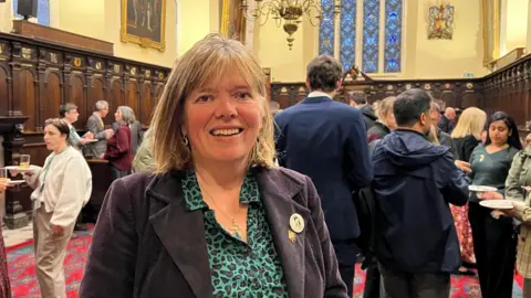 Diana Moore wearing a green and black shirt and a blue corduroy jacket with and Exeter badge on her lapel and another unknown badge with people in the background at the Guildhall