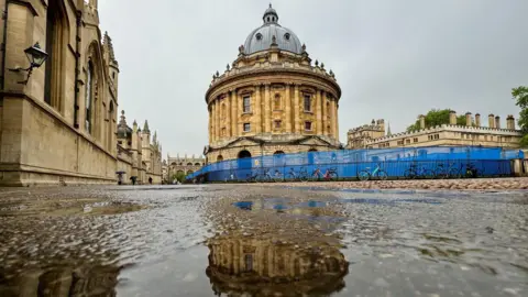 WEDNESDAY - A damp view of the Radcliffe Camera in Oxford with a grey sky behind it and its reflection in a puddle in the foreground