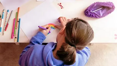 A stock picture taken from above of a girl with a purple jumper drawing a rainbow. Her face is not shown and crayons sit on her left hand side along with a purple pencil case on her right. 