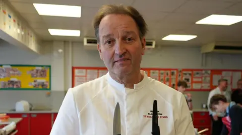Galton Blackiston, a chef who is standing inside a food technology kitchen classroom. He is in chef whites and is looking directly at the camera, and appears to be holding two knives.