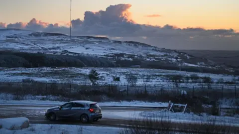 Getty Images A frosty mountain landscape. A single car is driving along a main road and there are visible signs of car tracks in the frost. The sun is beginning to rise from behind the clouds and a large number of fields are white with frost 