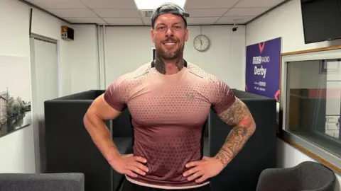 A man in a peach t-shirt with large arm muscles with a cap on backwards.