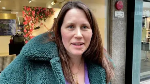 Robby West/BBC Rachel Martin, a woman looking at the camera and smiling. She has brown hair and is wearing a blue fluffy coat and purple top with gold necklaces. She is stood in front of a shop window. 