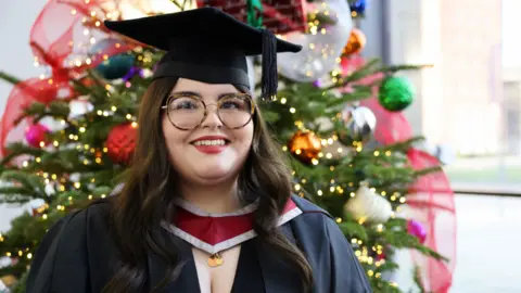 Swansea University Ayla Jones, a white young woman with long brown slightly wavy hair. She has brown eyes and wears large tortoiseshell-framed glasses. She has dark red lipstick on and is dressed in a graduate's cap and gown, with a red and grey-edged piece at the front of her neck. She is standing in front of an out of focus brightly decorated Christmas tree. She is smiling and looking slightly to the right of the camera.