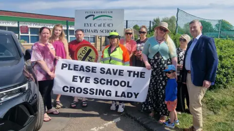 Protesters standing in front of Eye Primary School holding a white banner with black writing saying "please save the Eye school crossing lady".