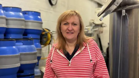 Head and shoulder shot of Alison Davis  with shoulder-length fair hair and wearing a pink and white striped hooded top. To her right is a high stack of metal beer containers.