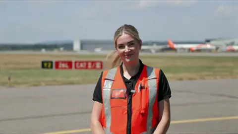 Danyell stands on the side of the runway in an orange high vis jacket. Planes are in the background.