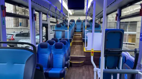 The inside of one of the new Vectio C buses, it has blue seats, with a wooden floor. There are white bars to hold on to and a yellow trim on the floor. 